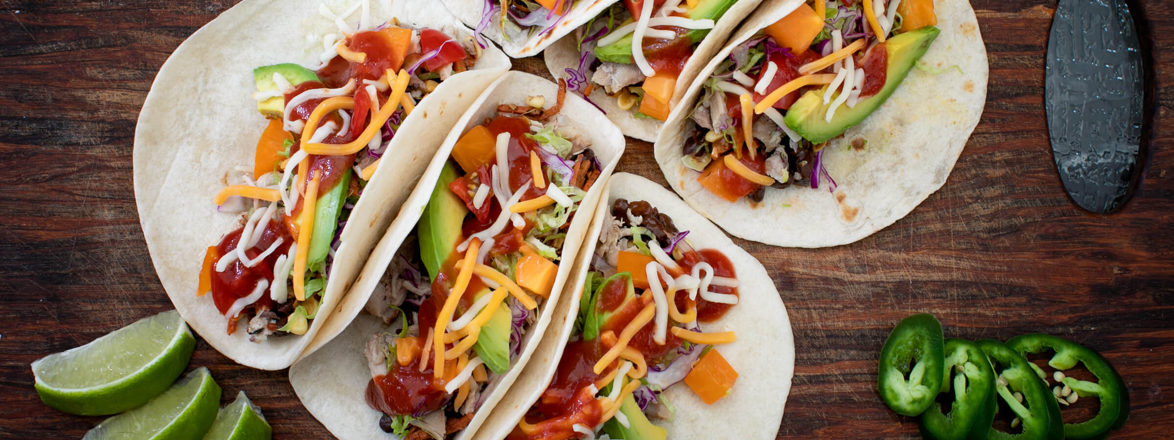 A series of tacos in flour tortillas on a cutting board with jalepenos and sliced limes
